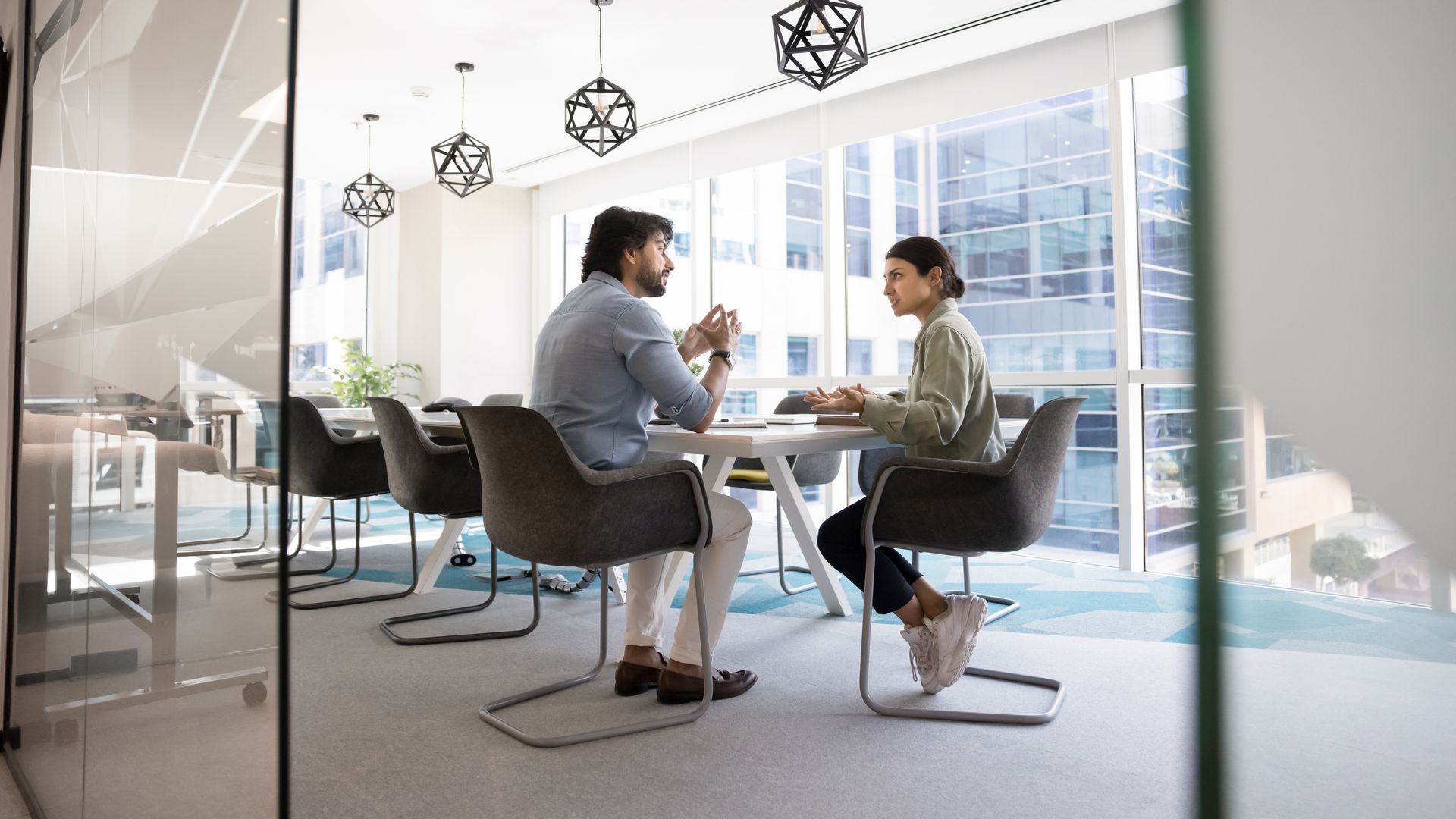 Two people sitting at a table in a modern, glass-walled office having a conversation.