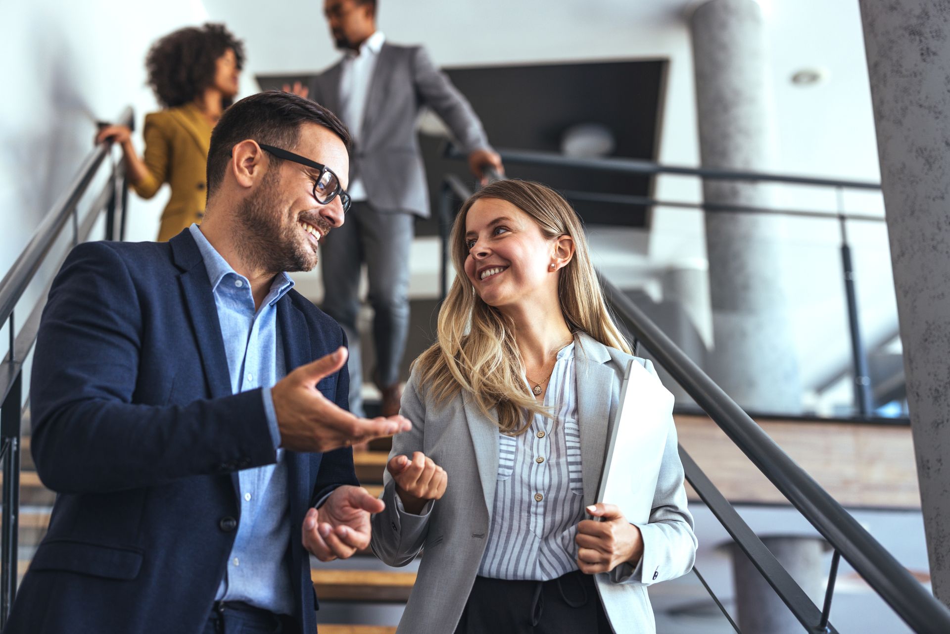 Business colleagues in formal attire smiling and talking while walking down a staircase in a modern office building.