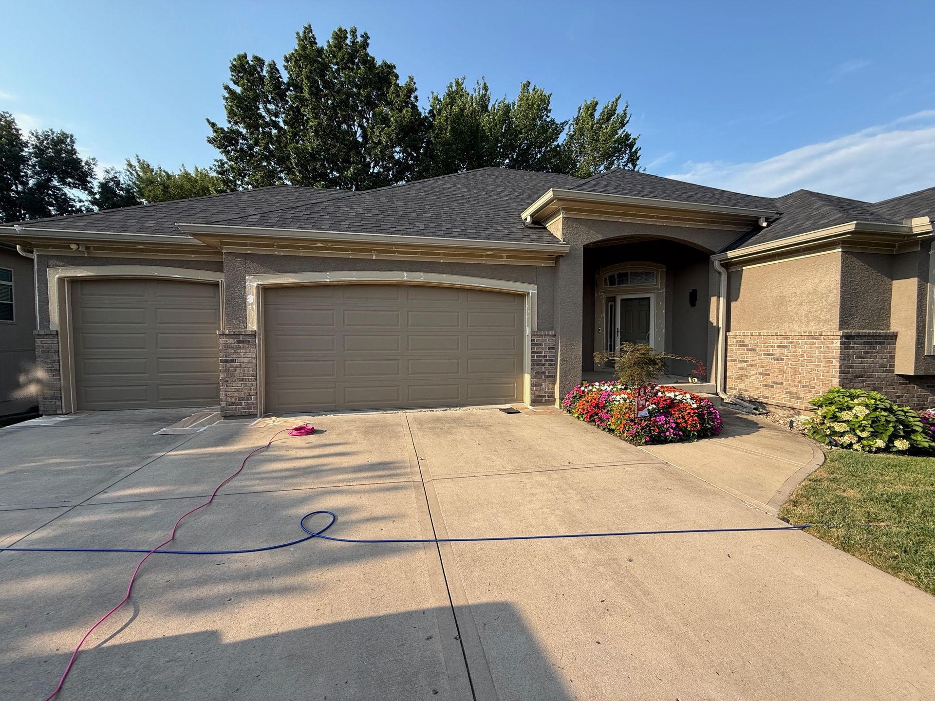 Tan house with a two-car garage and brown roof, flowers, and a driveway.