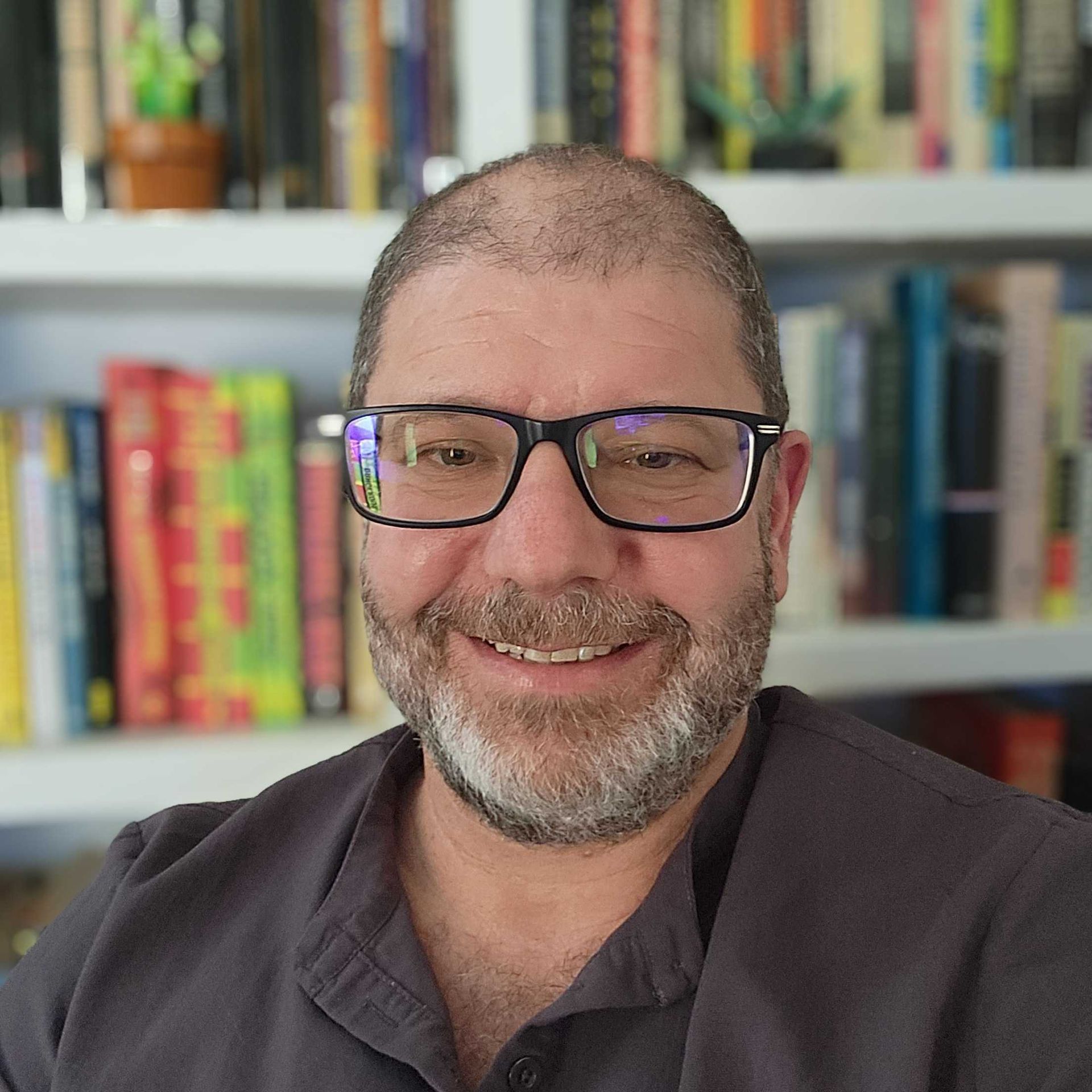 A man with glasses and a beard is smiling in front of a bookshelf.