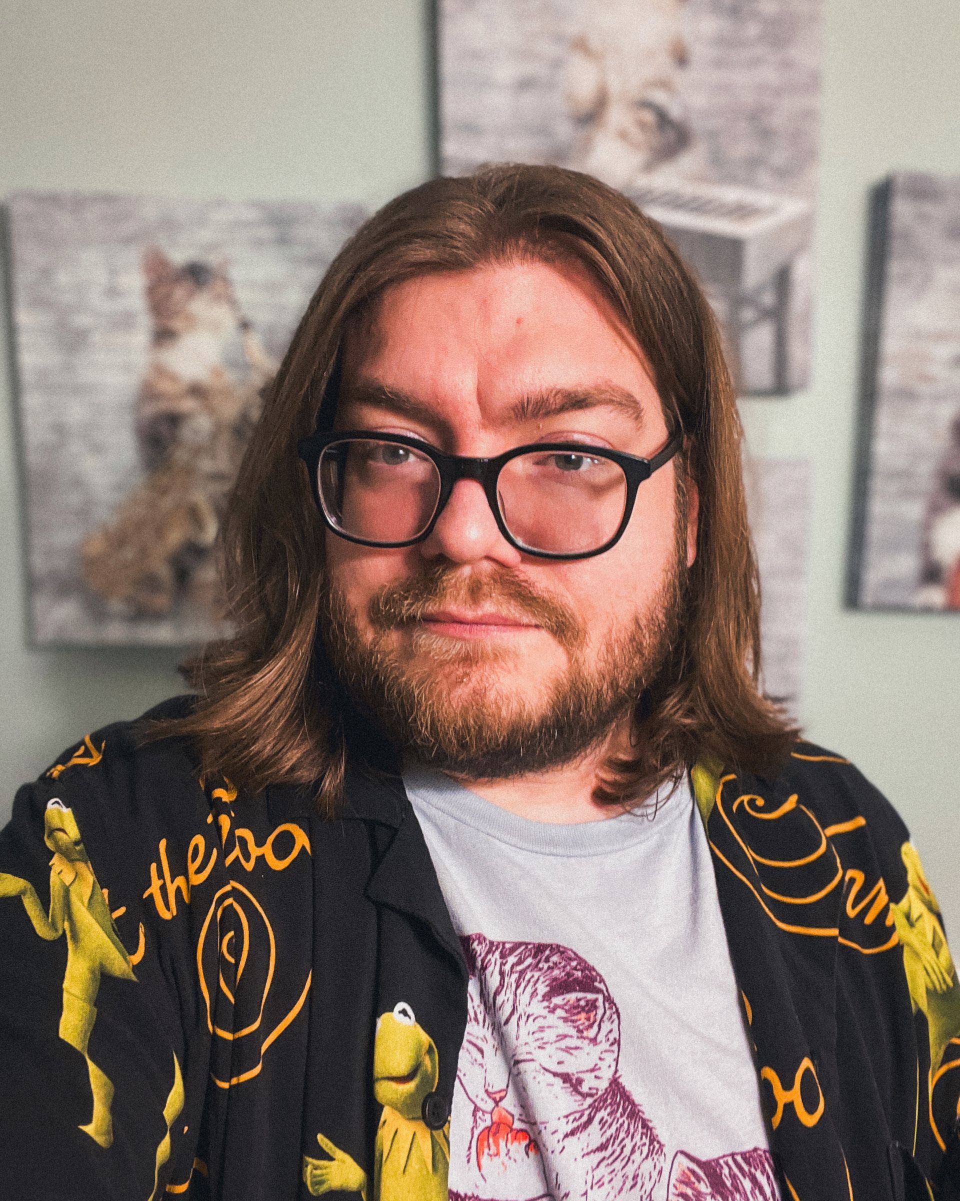 A man with glasses and a beard is standing in front of a wall with pictures on it.