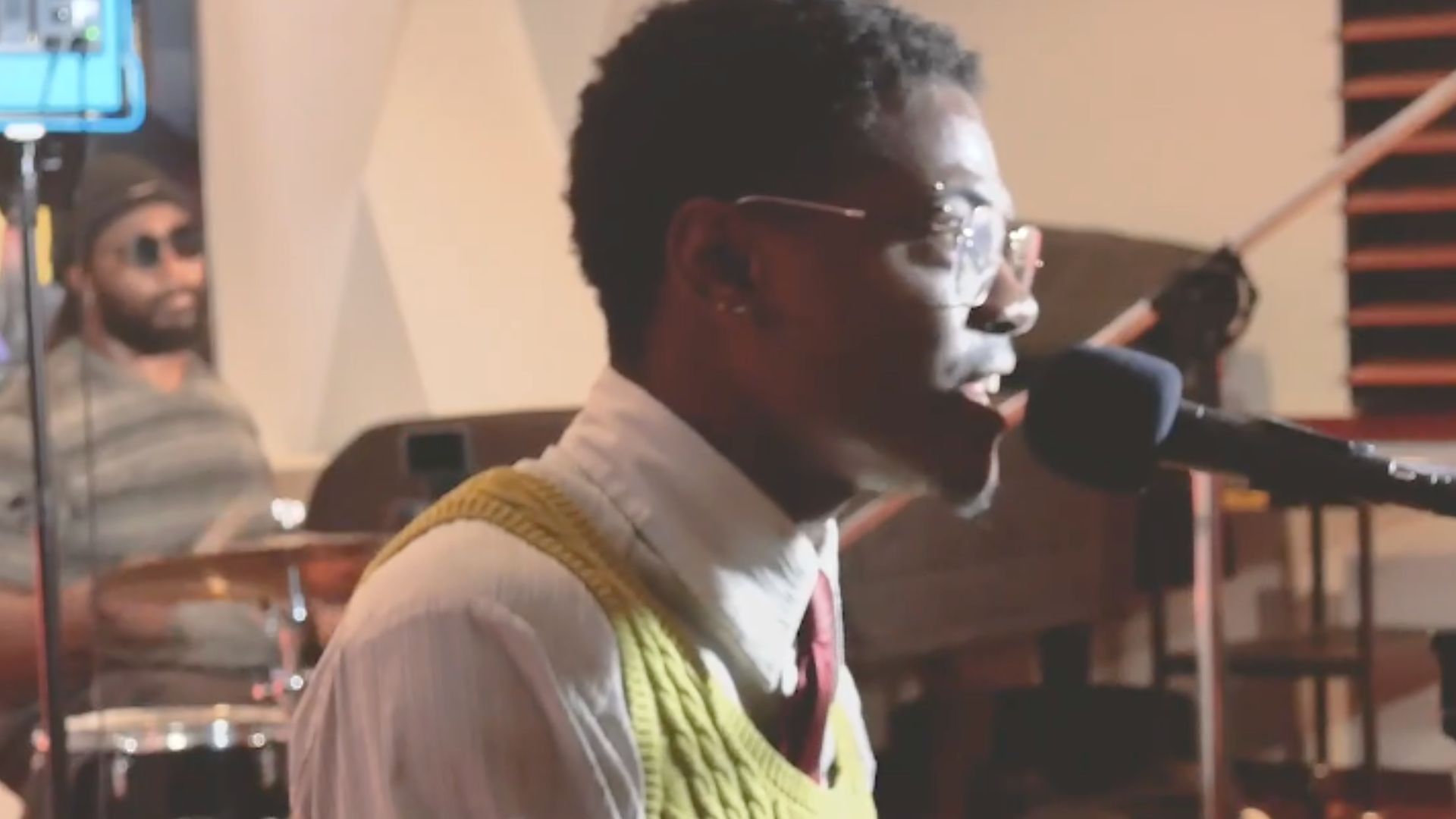Man singing into microphone at piano, wearing glasses, yellow vest, and shirt. Drummer in background.