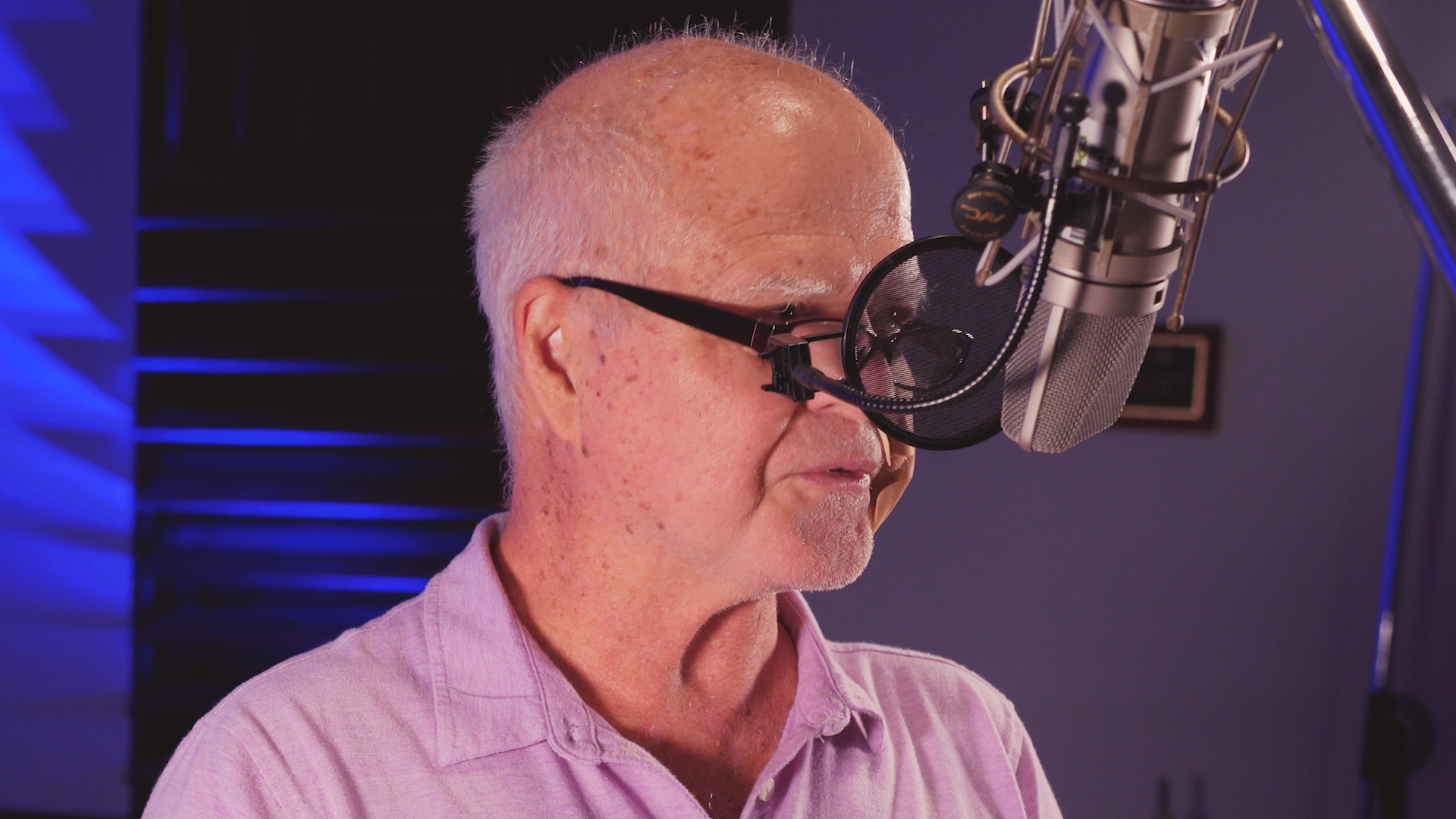 An older man wearing glasses is singing into a microphone.