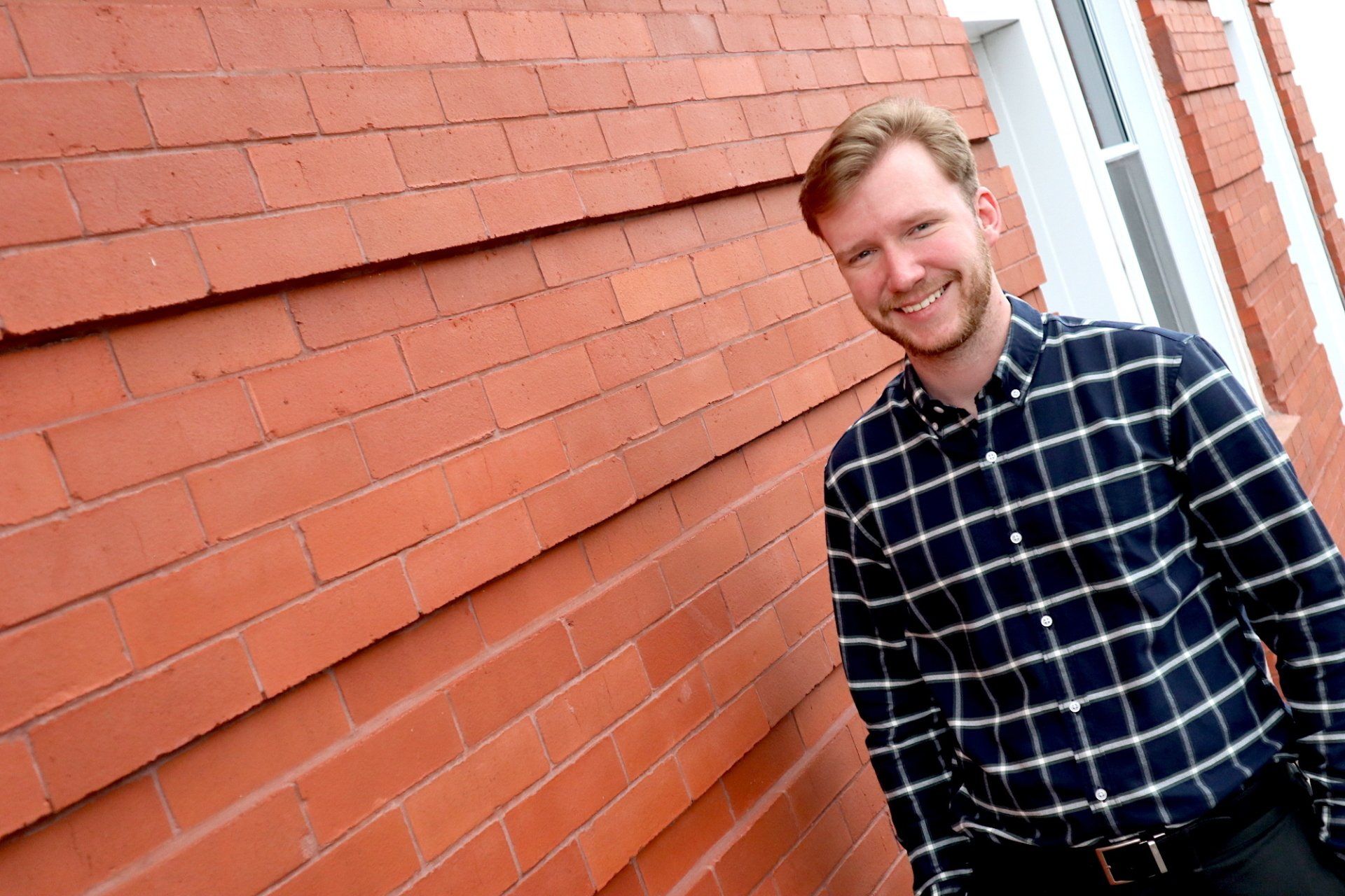 A man in a plaid shirt is standing in front of a red brick wall.