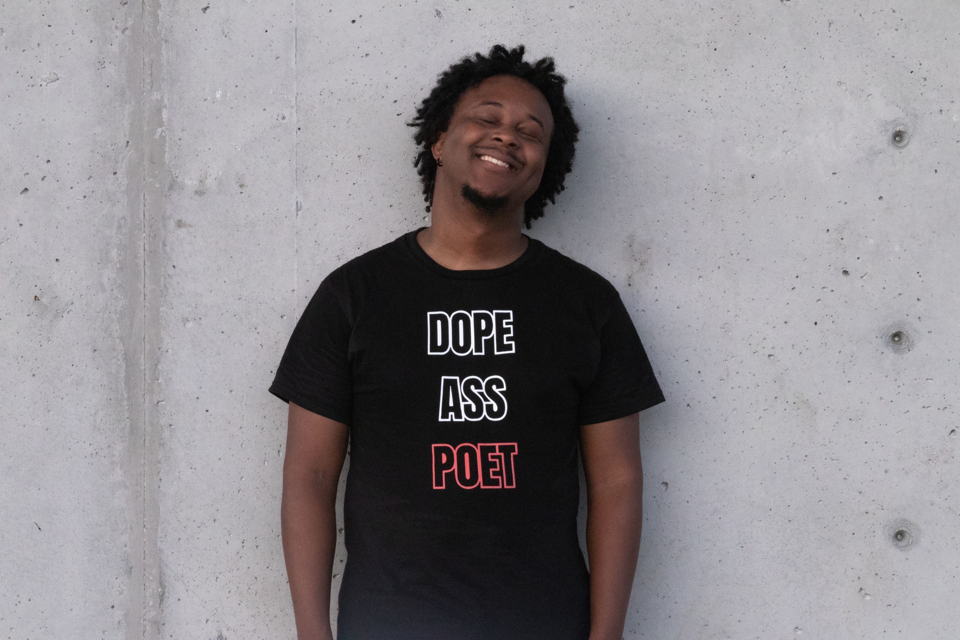 A person standing in front of a concrete wall smiling while wearing a t-shirt that says 
