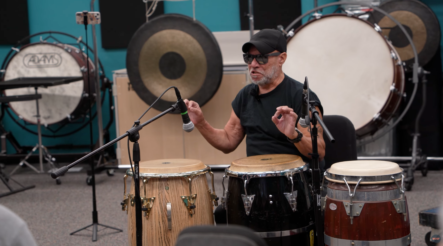 Man with glasses gesturing, seated at conga drums in a music studio, other percussion instruments visible.