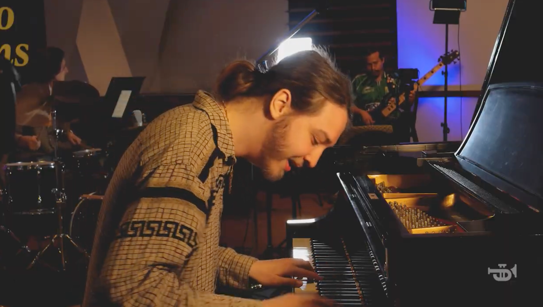 Pianist with long hair plays piano in a band. Other musicians in background: drums, bass, saxophone. Indoors, dim lighting.