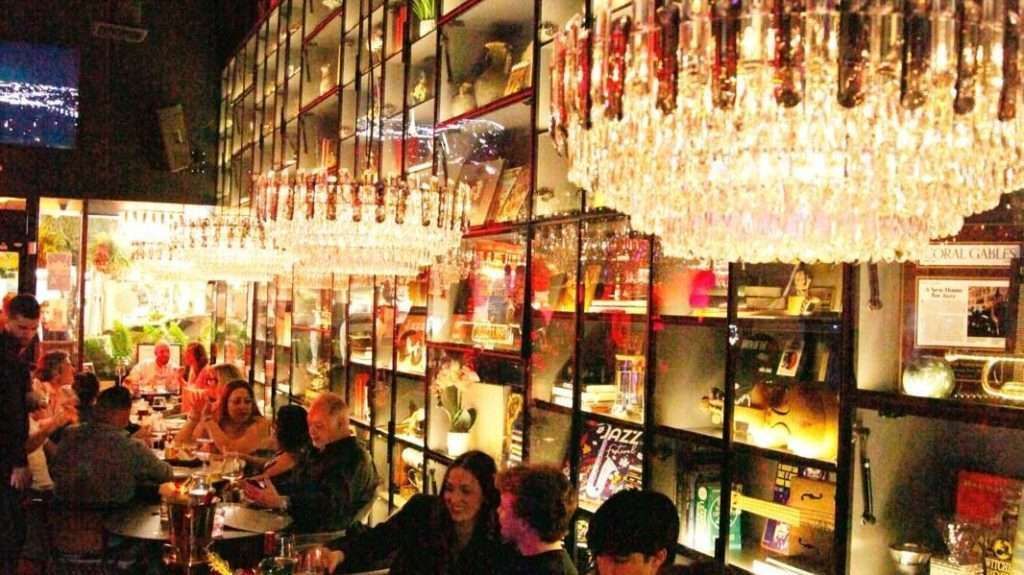 Bar interior, people at tables, long bar with shelves of objects, crystal chandeliers.