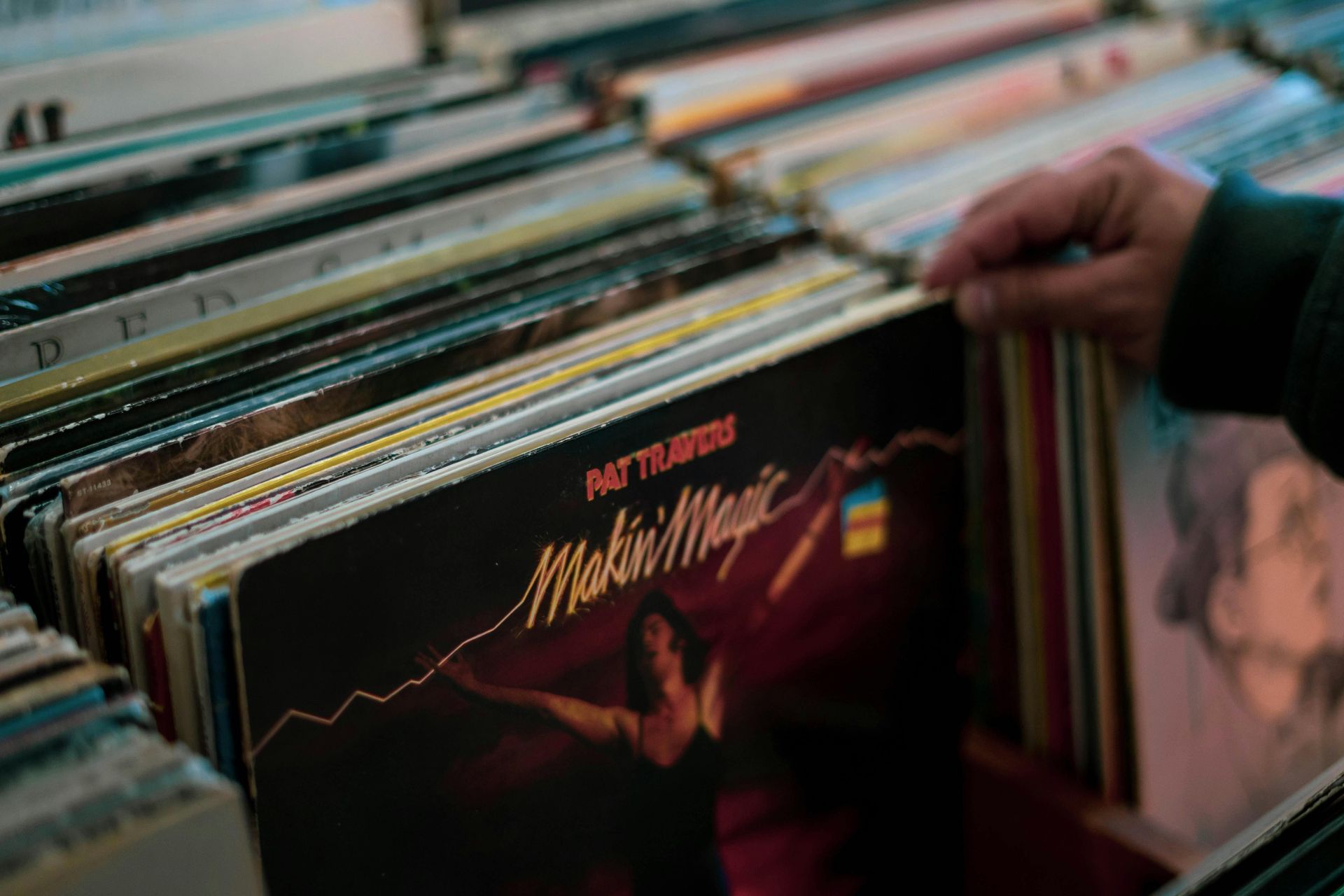Person's hand browsing vinyl records in a record store. Albums are in rows, and one cover is visible: