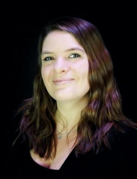 A woman with long brown hair is smiling in front of a black background.