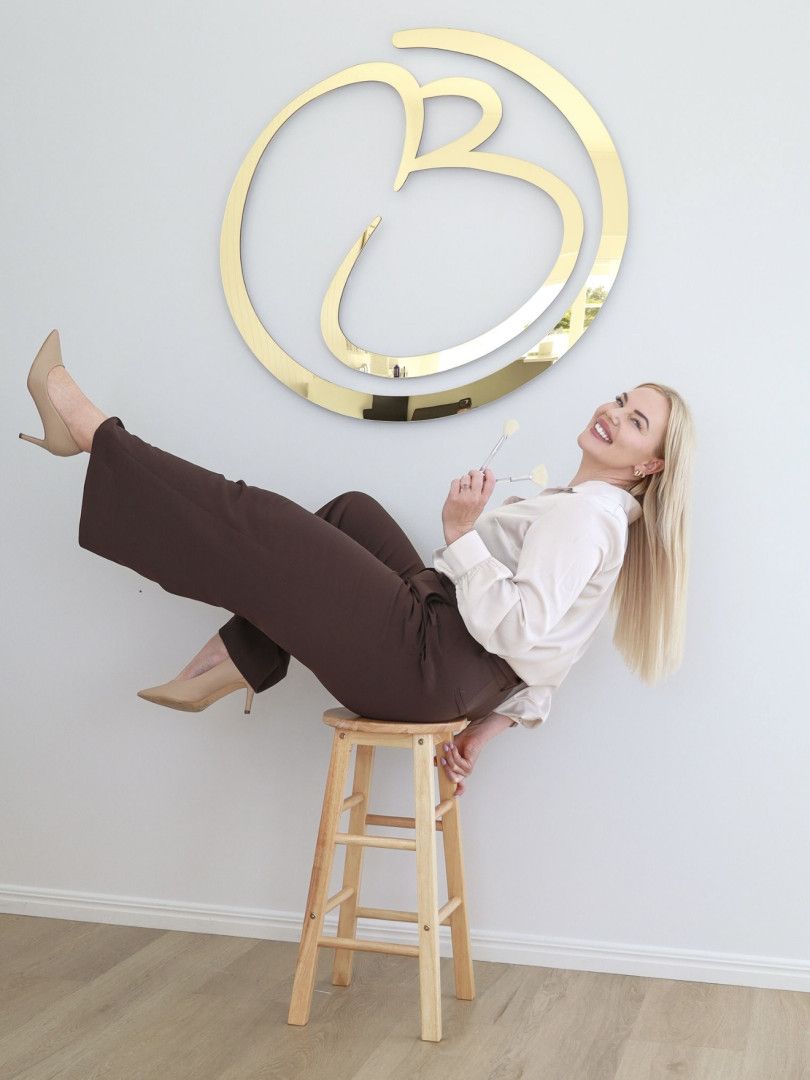 Woman in brown pants and heels laughing while perched on a stool under a gold logo.