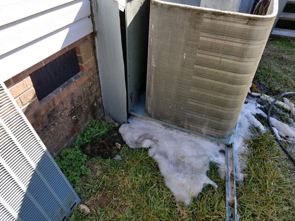 A dirty air conditioner is sitting in the grass next to a brick wall.