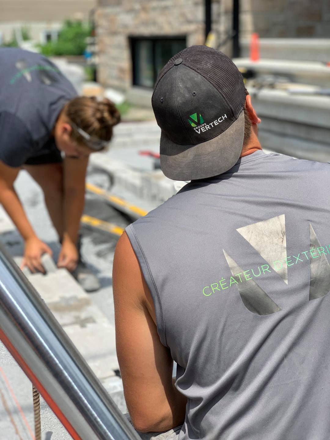 Deux ouvriers en chemise grise posent du pavage à l'extérieur. L'un porte un chapeau ; l'autre travaille avec des lunettes de sécurité.