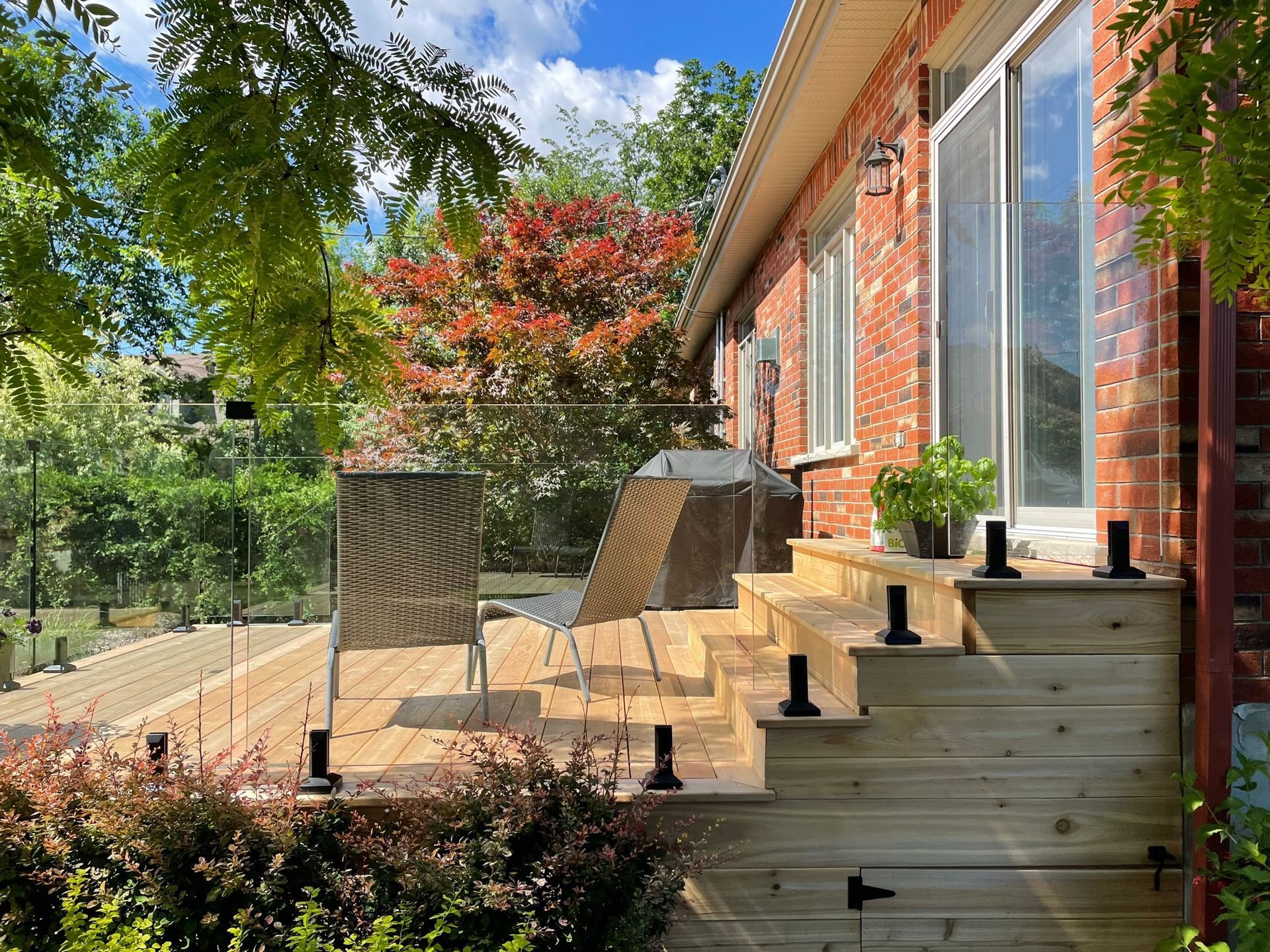 Terrasse en bois avec garde-corps en verre, escalier menant à une maison en briques. Jardins et arbres alentour.