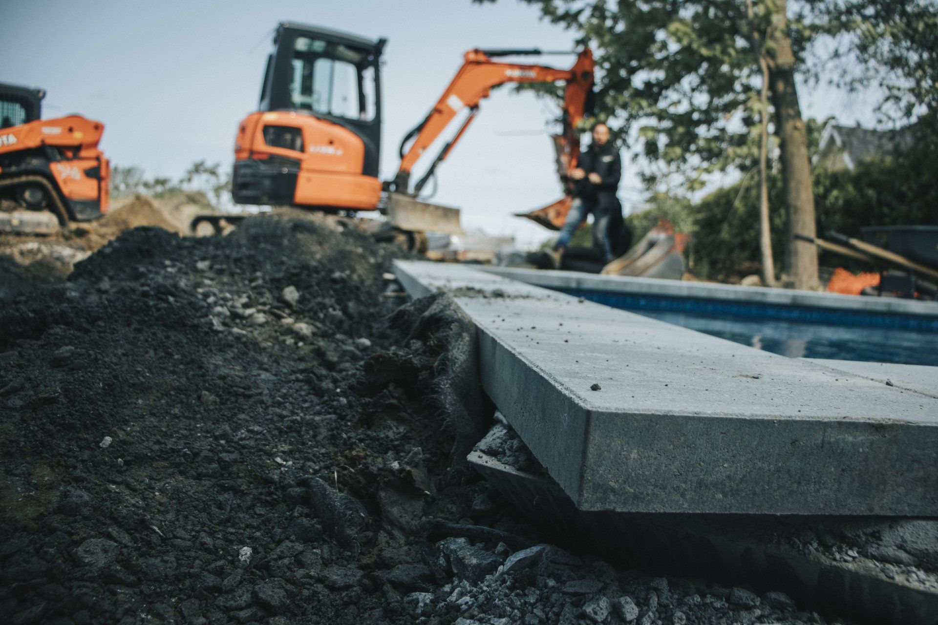 Chantier de construction avec une bordure en pierre autour d'une piscine. Une pelleteuse et une personne sont visibles à l'arrière-plan.