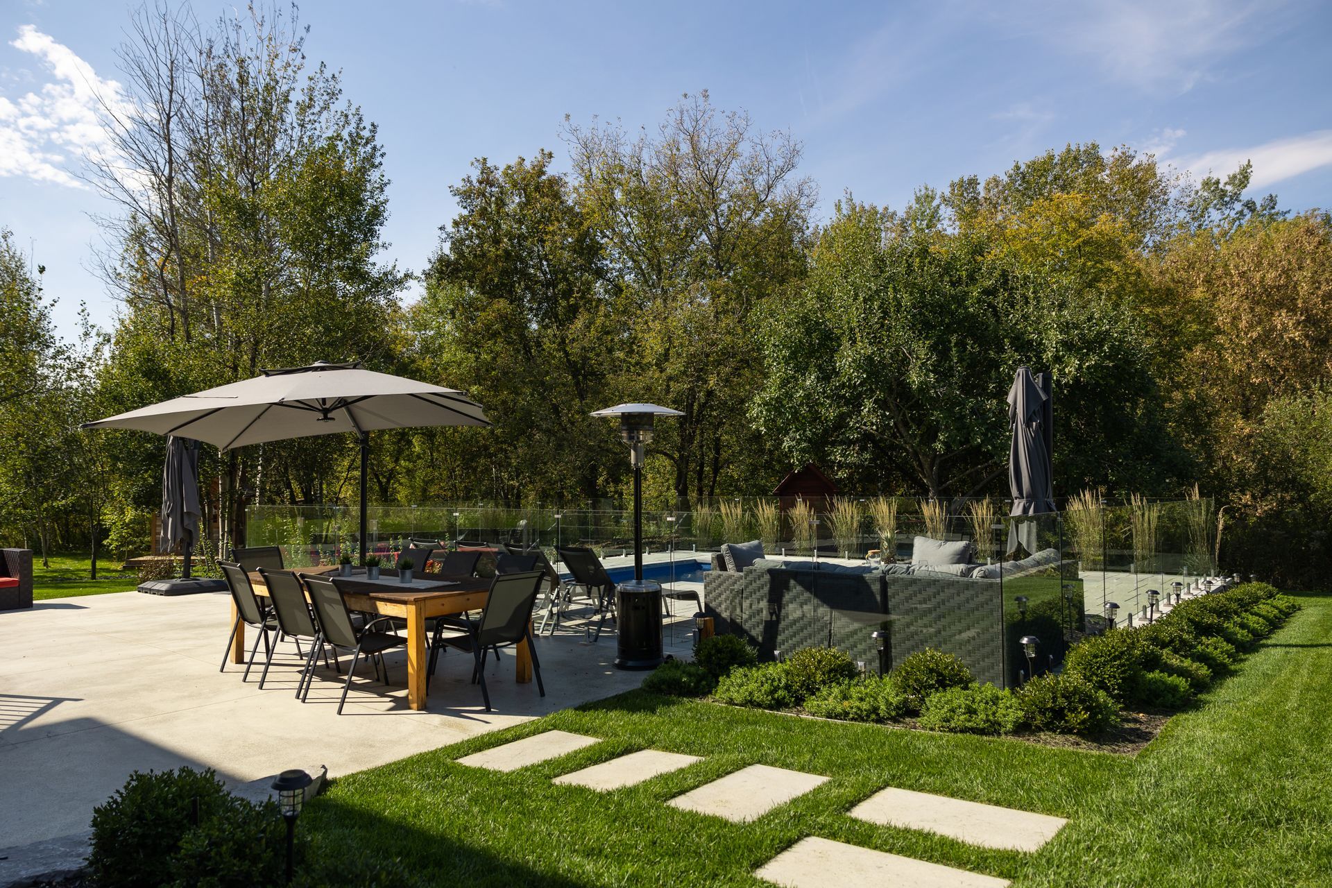 Terrasse avec table et chaises sous un parasol, au bord d'une piscine. Pelouse verte, arbres et ciel bleu en arrière-plan.