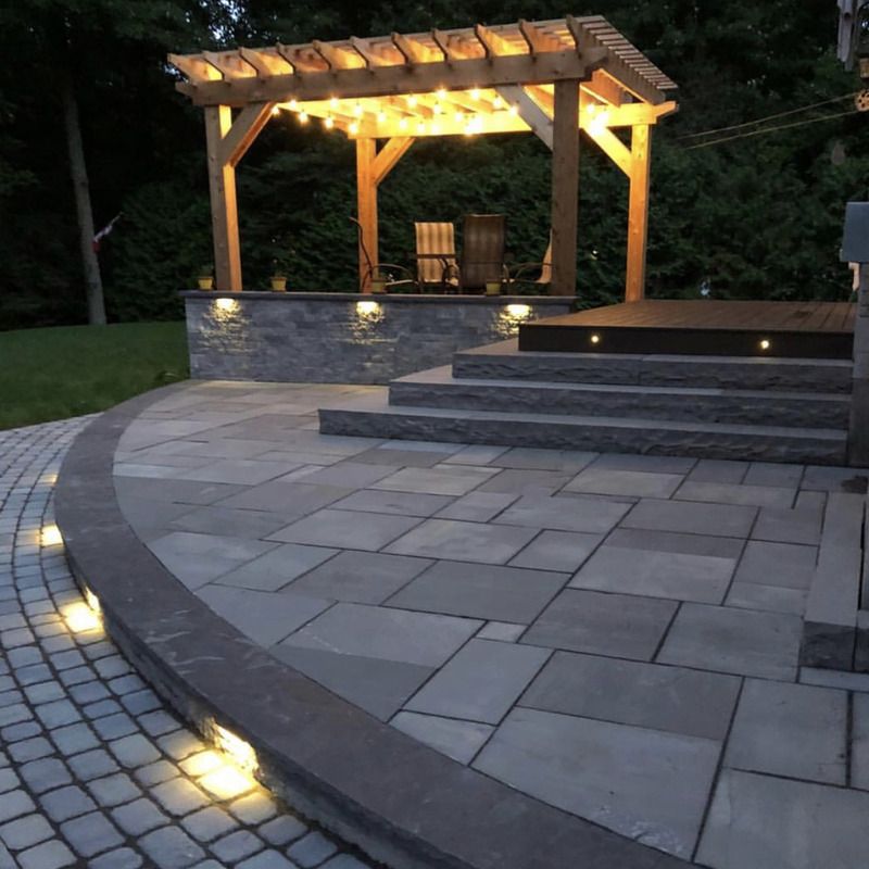 Une terrasse en pierre avec une pergola éclairée par des guirlandes lumineuses. Un escalier mène à la pergola, avec un éclairage d'appoint.