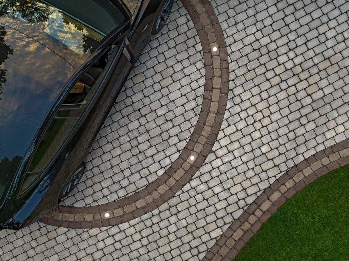 Voiture noire garée sur une allée pavée de briques avec une bordure brune incurvée et de petites lumières ; pelouse verte adjacente.
