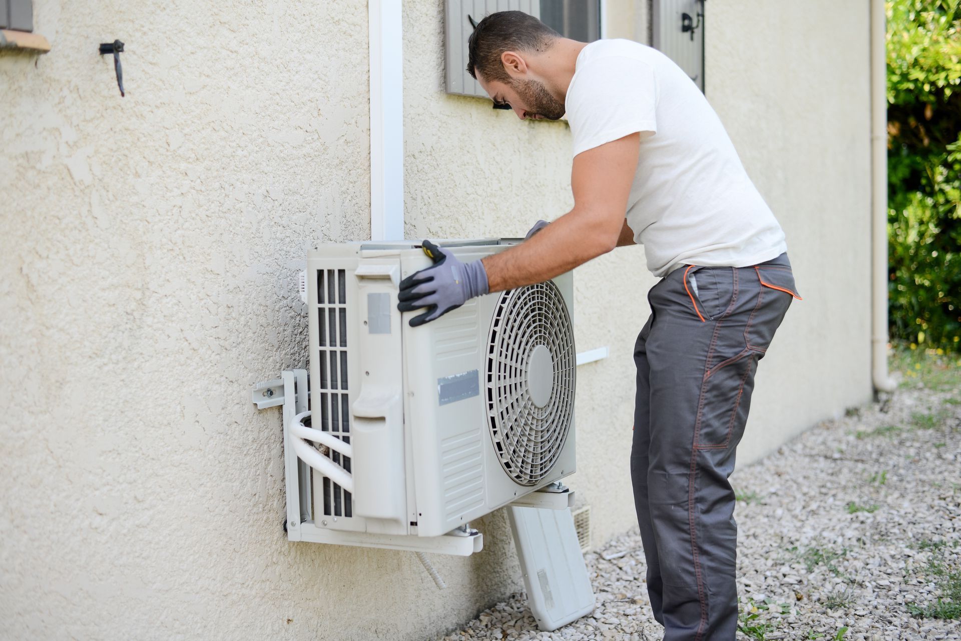 Un homme en pantalon gris et chemise blanche installe une unité de climatisation à l'extérieur.