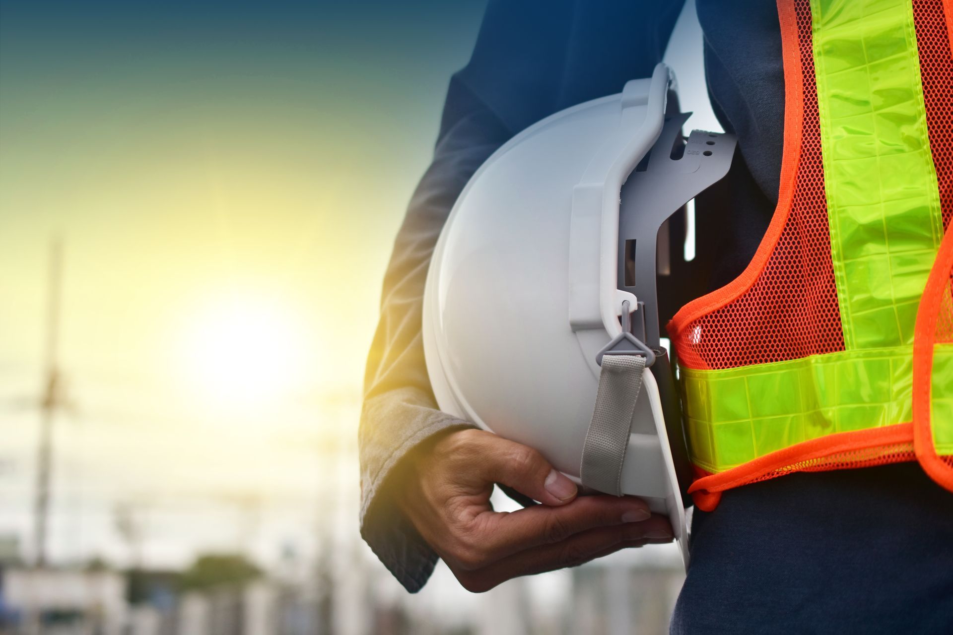 Ouvrier tenant un casque blanc à côté d'un gilet de sécurité fluo sur un chantier de construction au coucher du soleil