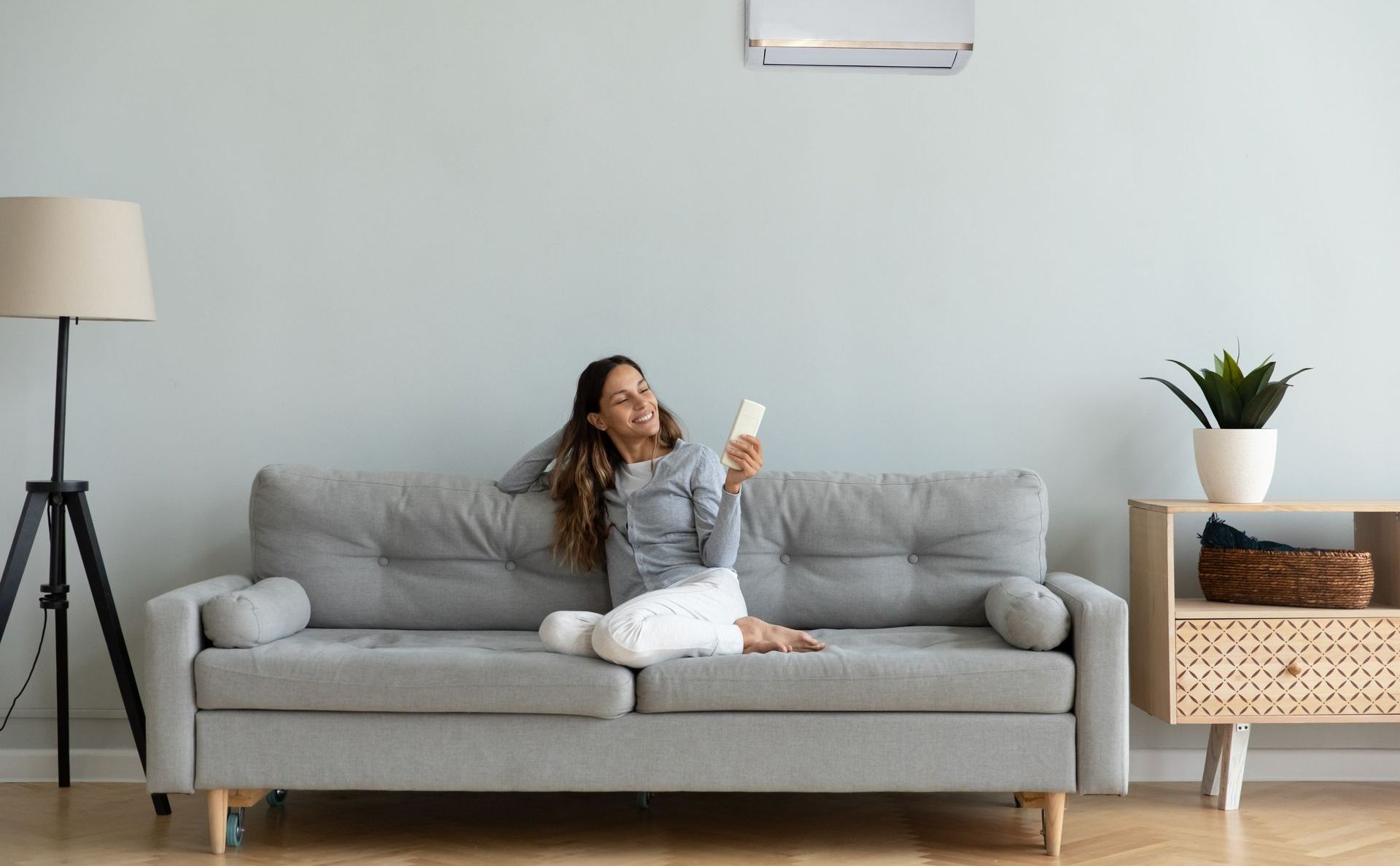 Femme assise sur un canapé gris, utilisant une télécommande, avec un climatiseur au-dessus, dans un salon.