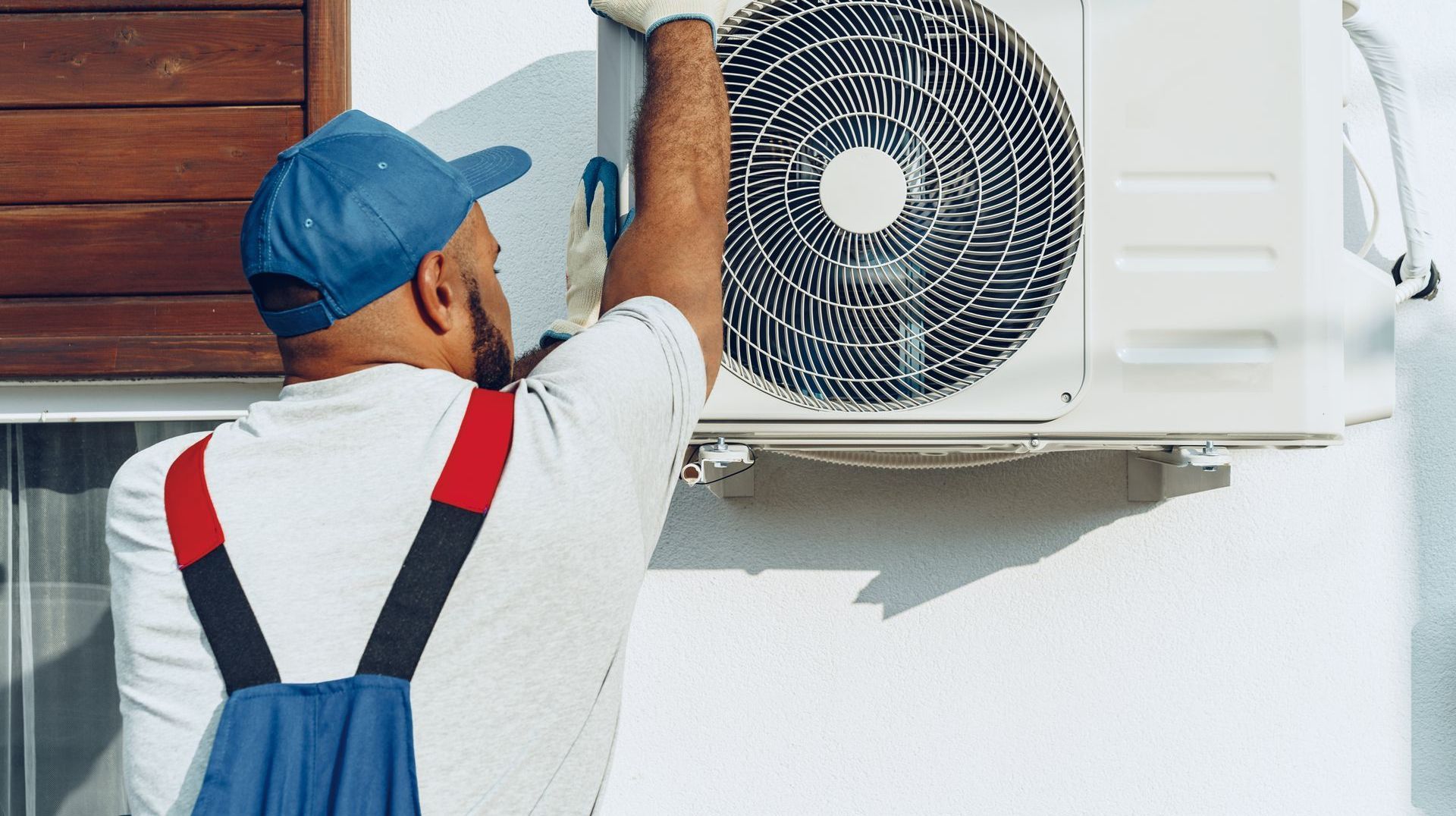 Un homme en salopette installe un climatiseur sur un mur extérieur.