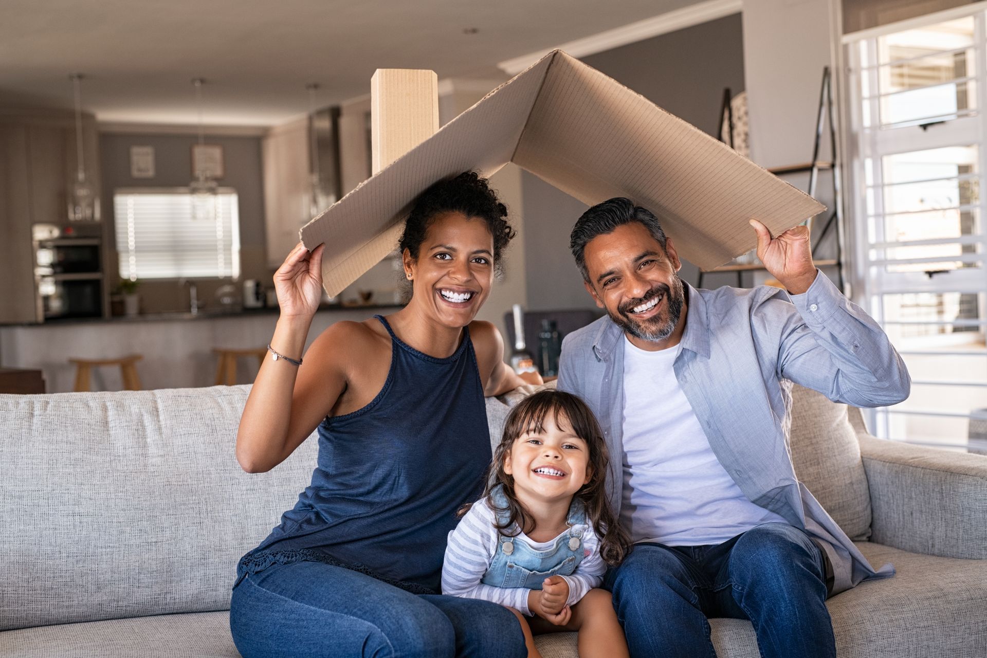 Une famille assise sur un canapé, souriante, tenant une boîte en carton comme un toit de maison.