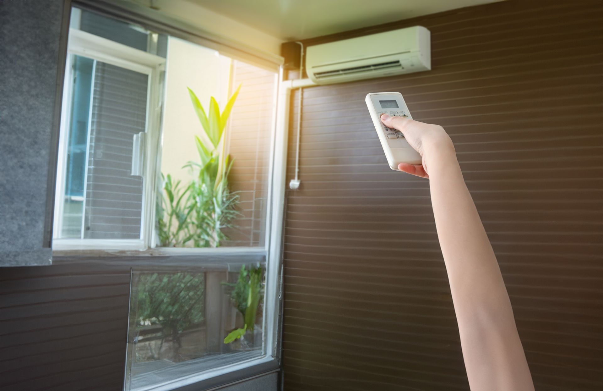 Le bras d'une personne tendant la main vers une télécommande de climatisation, avec un climatiseur sur le mur près d'une fenêtre.