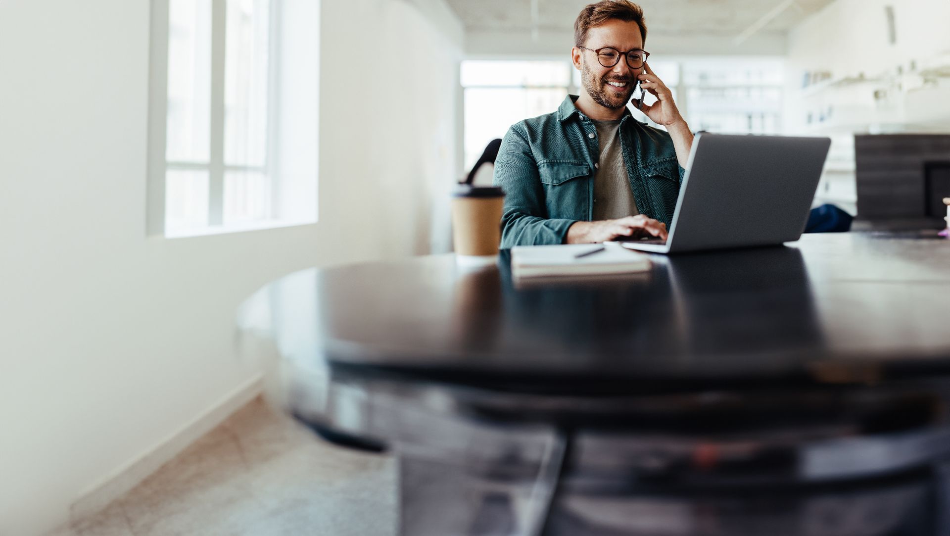 Un homme souriant au téléphone et tapant sur un ordinateur portable à une table ronde dans un bureau lumineux.