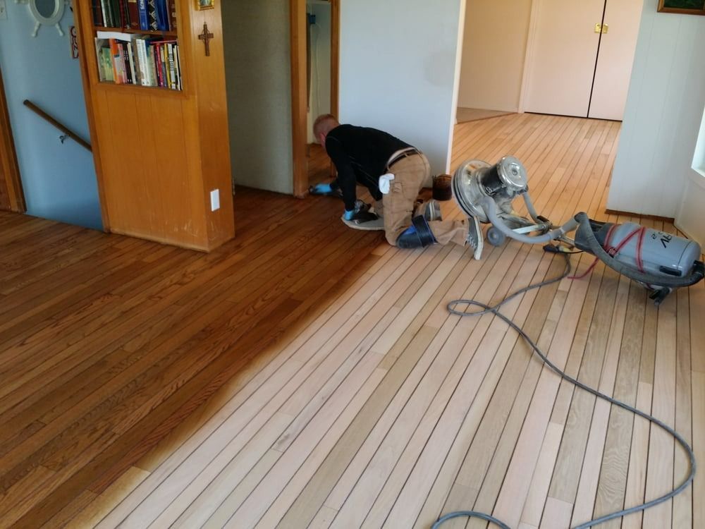 Man refinishing hardwood floor, using sanding machine and finishing. Light wood, half-stained brown.