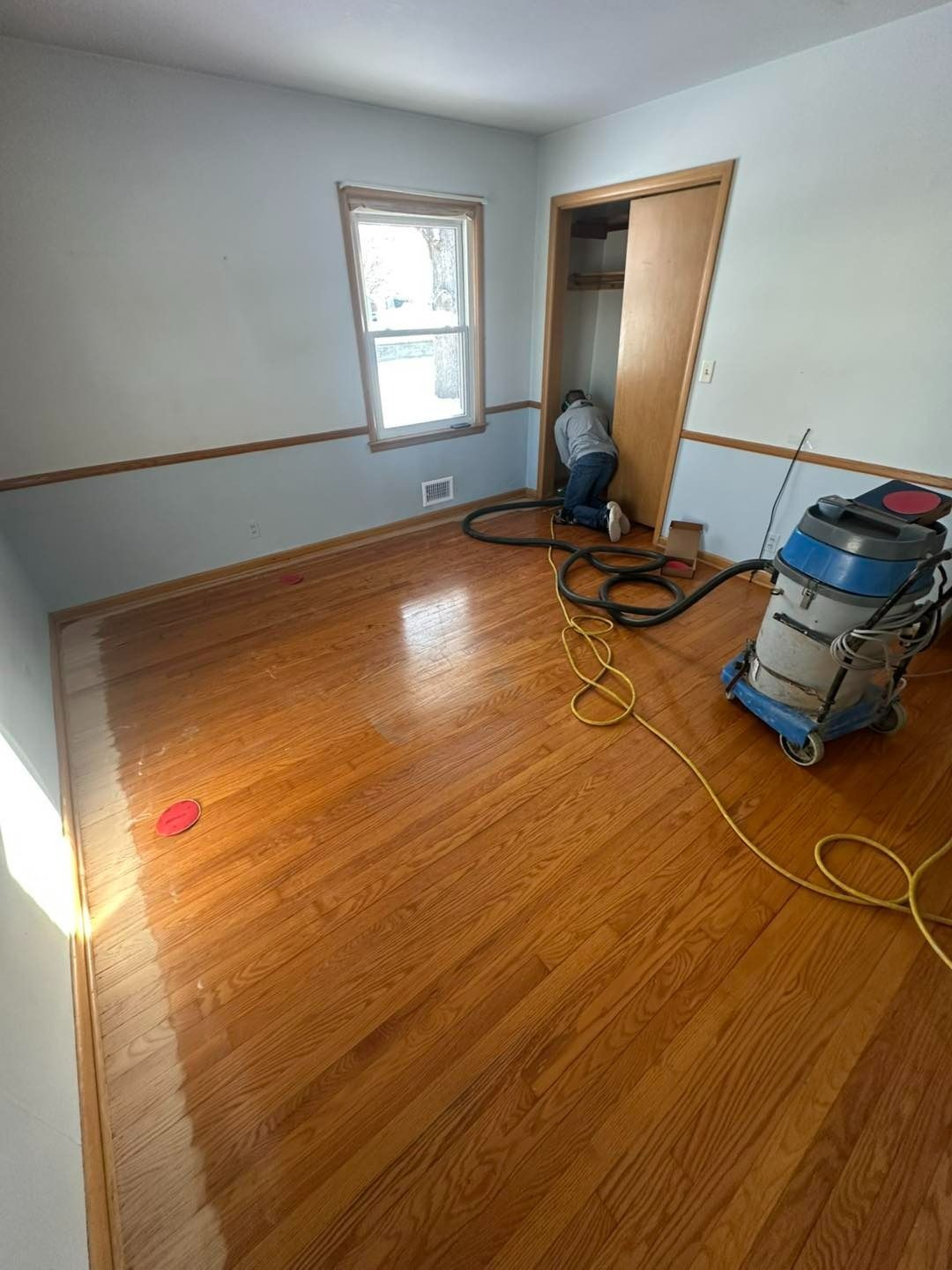 Person refinishing hardwood floor in a room with blue and tan walls, a window, and a closet.