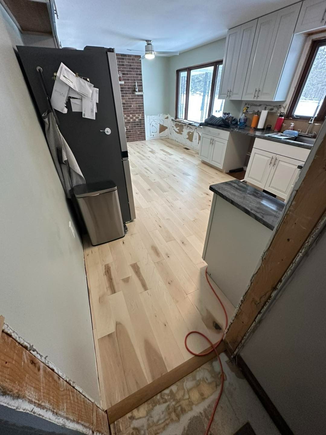 Kitchen renovation in progress: new light wood flooring, white cabinets, and a partially demoed wall.