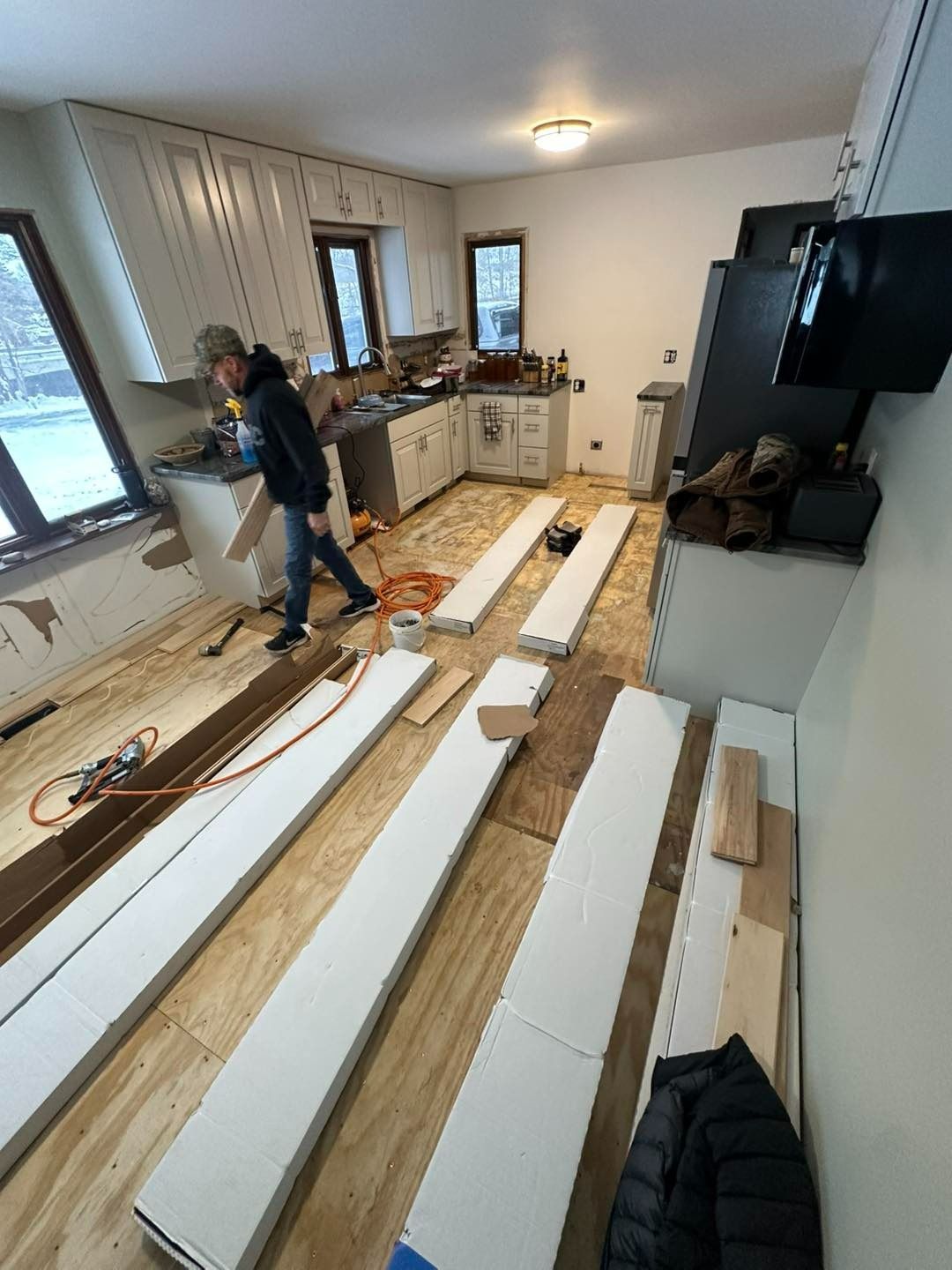 Person cutting wood in a kitchen. Flooring renovation in progress with exposed subfloor, cabinets, and appliances.