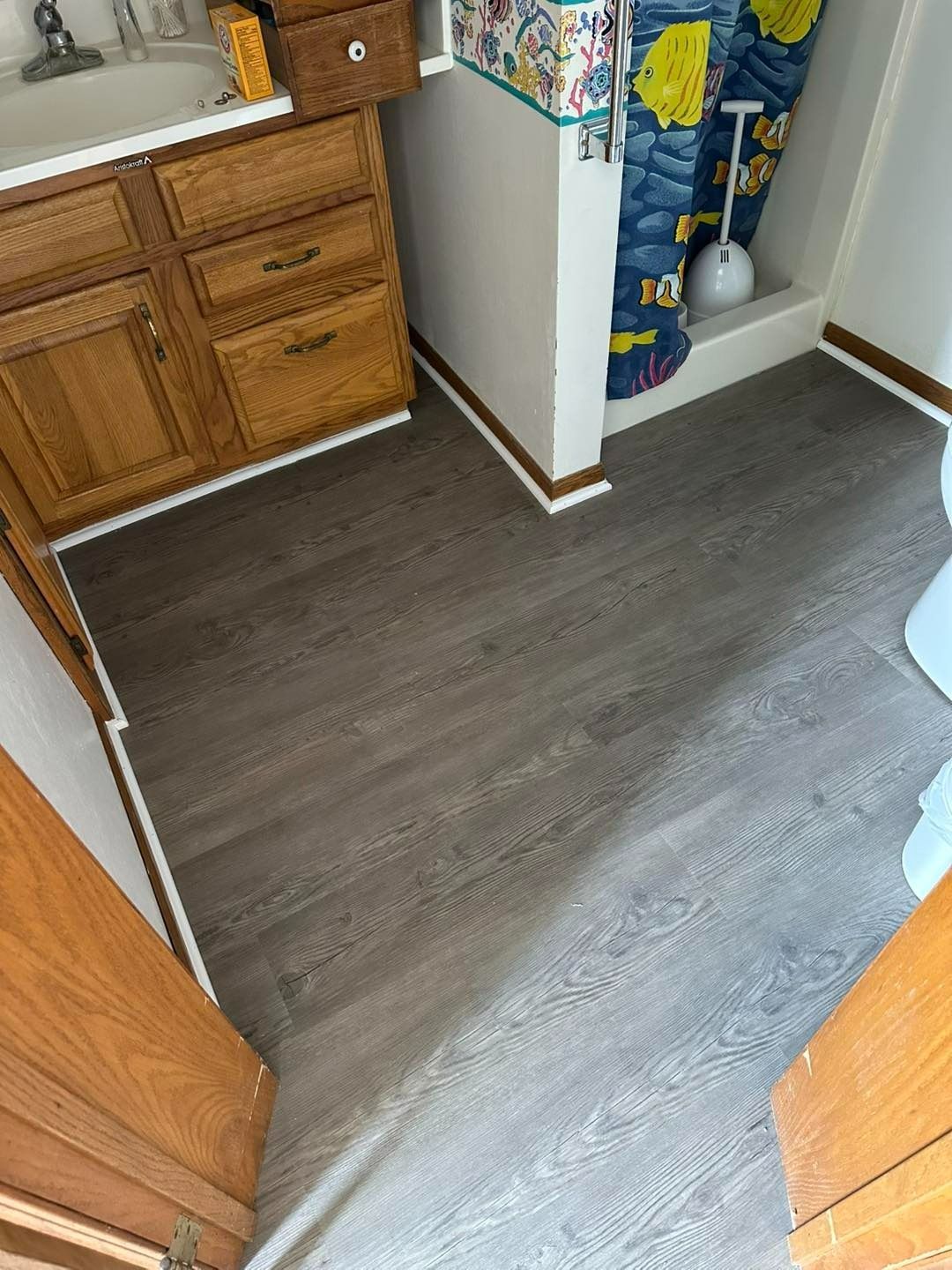 Bathroom with gray wood-look flooring, wood cabinets, and a blue and yellow shower curtain.