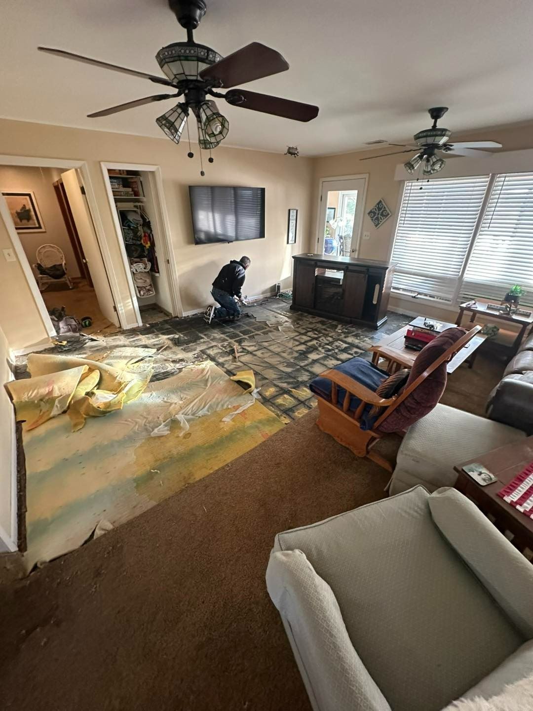 A person removes flooring in a living room. Damaged carpet, furniture, and debris are scattered.