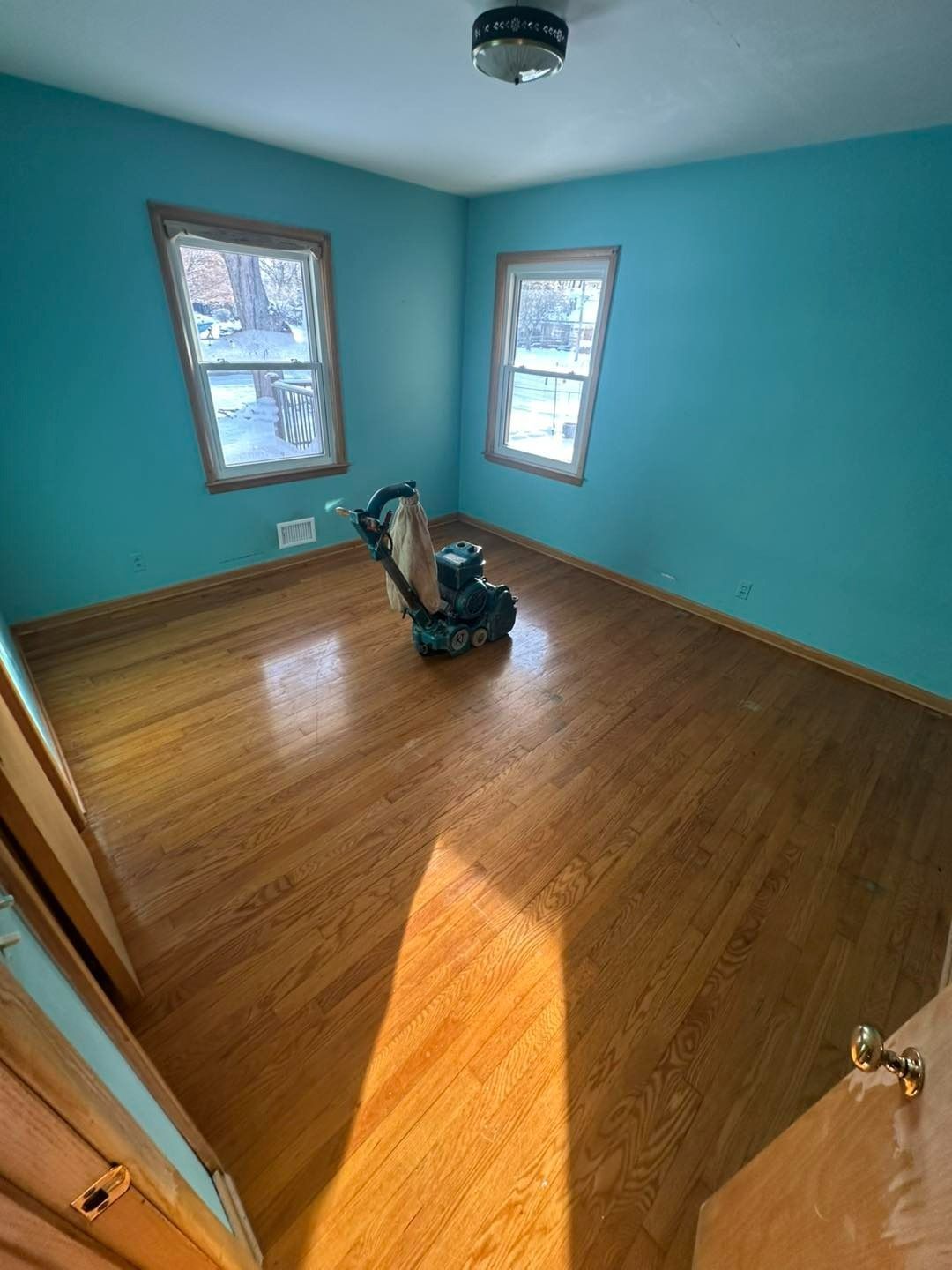 Room with wood floor being sanded; turquoise walls, windows, and sander visible.