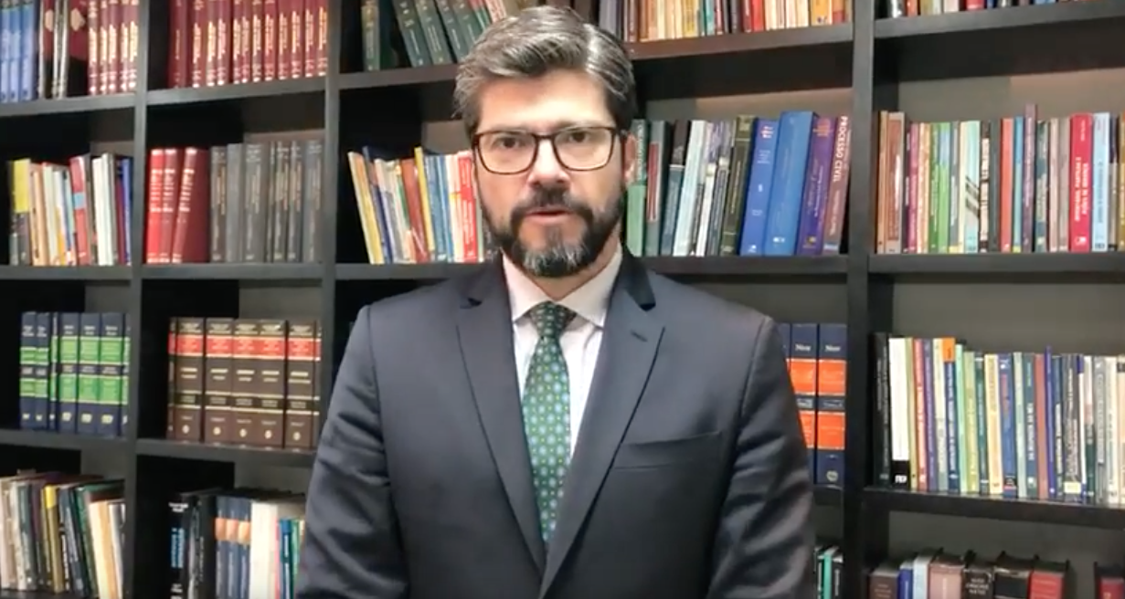 Um homem de terno e gravata está parado em frente a uma estante de livros.