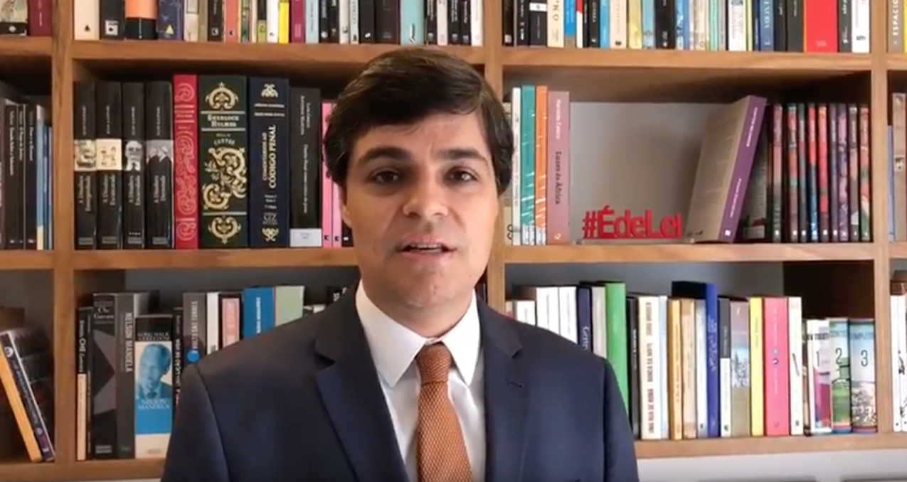 Um homem de terno e gravata está parado em frente a uma estante cheia de livros.