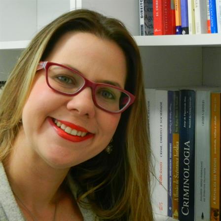 Uma mulher de óculos sorri em frente a uma estante cheia de livros, incluindo criminologia