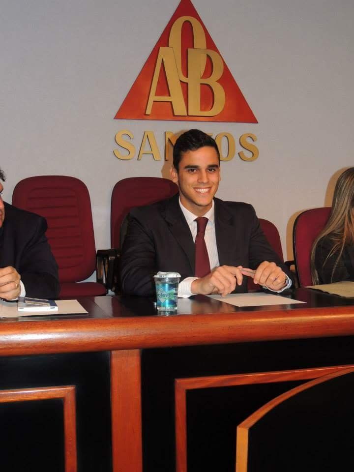 Um homem de terno e gravata está sentado em uma mesa em frente a uma placa que diz ab santos