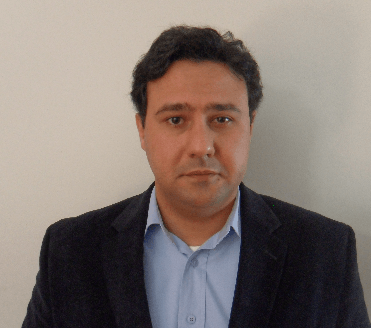 Um homem de terno e camisa azul fica em frente a uma parede branca