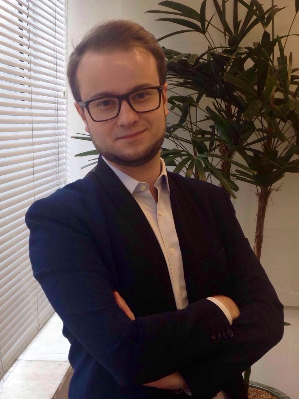 Um homem de terno e óculos está parado com os braços cruzados em frente a uma planta.