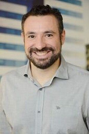 Um homem com barba está sorrindo para a câmera enquanto veste uma camisa cinza.