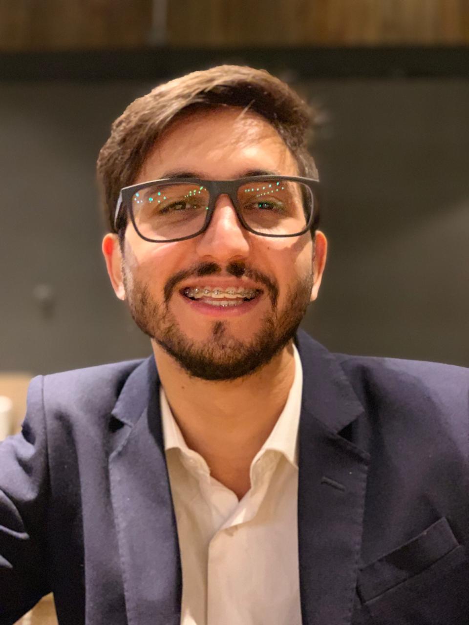 Um homem com barba usando óculos e aparelho está sorrindo para a câmera.