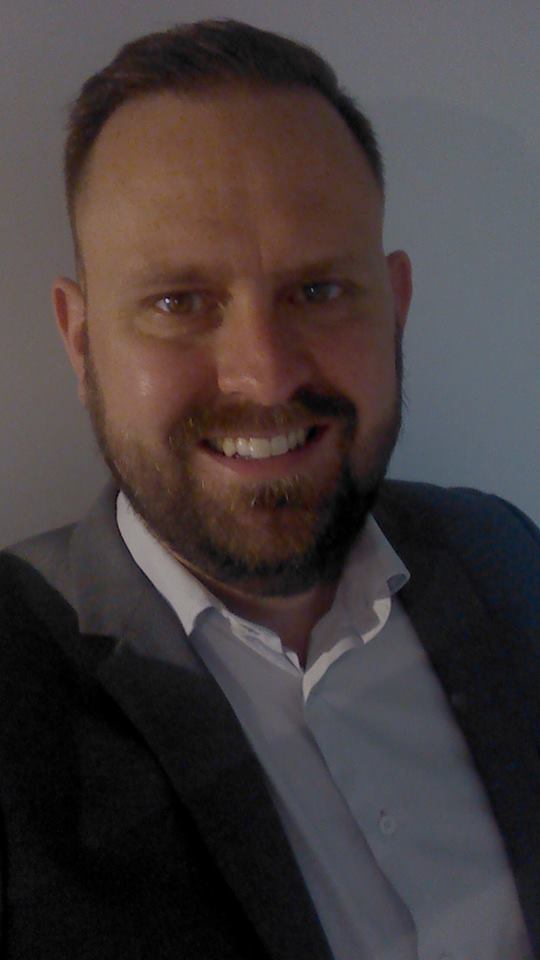 Um homem com barba está vestindo terno e sorrindo para a câmera.