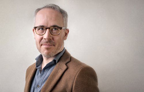 Um homem de óculos e jaqueta marrom está parado em frente a uma parede branca.