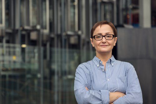 Uma mulher de óculos está parada com os braços cruzados em frente a um prédio.