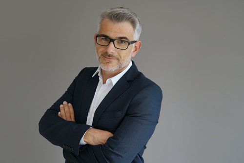 Um homem de terno e óculos está de pé com os braços cruzados.