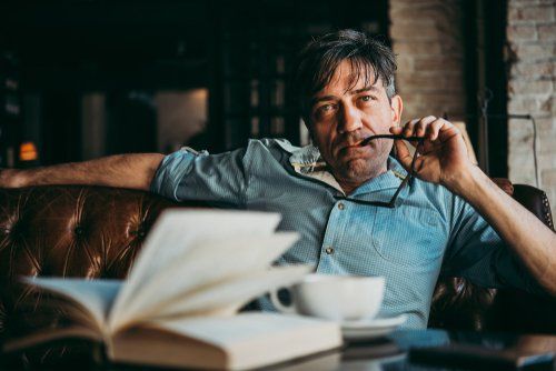 Um homem está sentado em um sofá lendo um livro e tomando café.