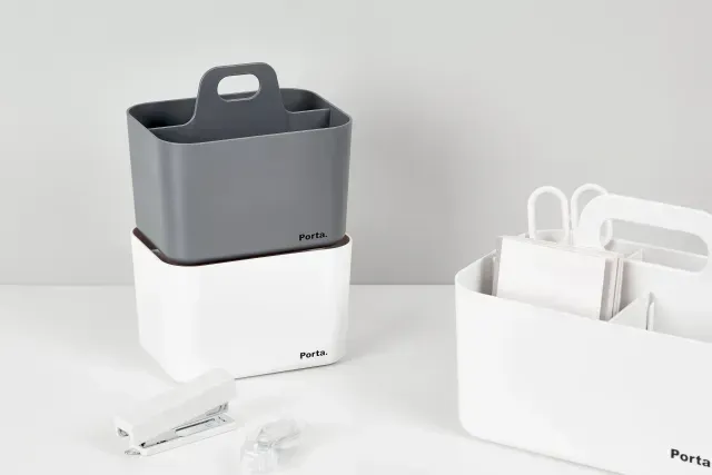 Un recipiente gris y blanco con asa está sobre una mesa junto a una grapadora y unas tijeras.